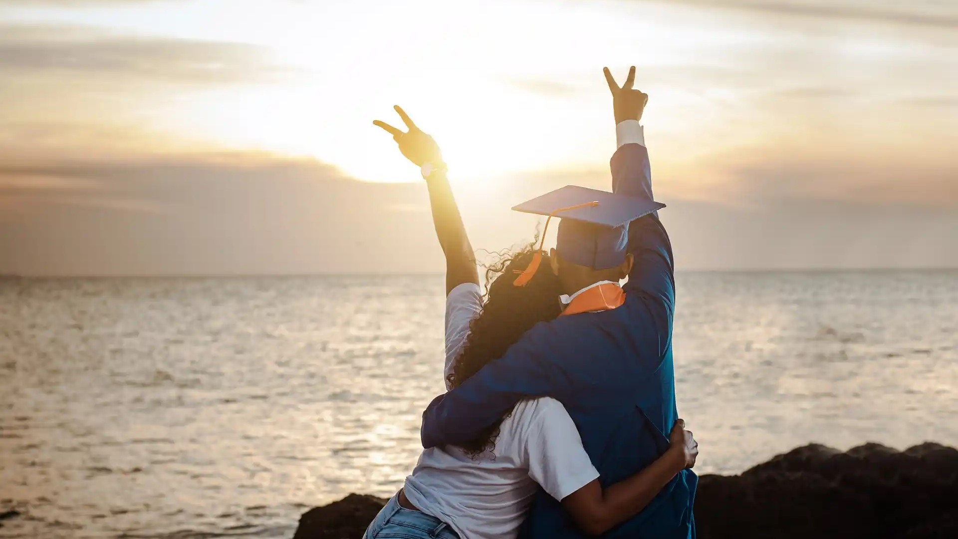 a man and woman in graduation cap and gown hugging and holding hands up